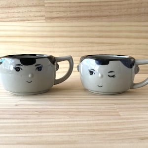 May include: Two ceramic mugs with painted faces. One mug has a closed eye and the other has an open eye. Both mugs have black hair and a light grey body.