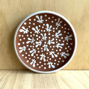 Puede incluir: Un plato pequeño y redondo de cerámica con un borde blanco y un interior marrón. El plato está decorado con un patrón geométrico blanco de puntos y flores estilizadas.