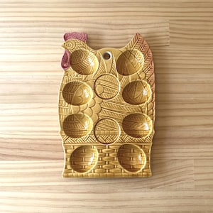 Pode incluir: Um porta-ovos de cerâmica em forma de galinha, com um esmalte amarelo-castanho. O suporte tem espaços para ovos e é decorado com um padrão de tijolos na base e uma cabeça de galo no topo. A crista é de cor vermelho-rosa.
