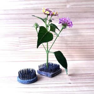 Op de afbeelding: Een bloemstuk met paarse en gele bloemen en groene bladeren, gerangschikt in twee blauwe kenzans. Eén kenzan is rond, de andere is vierkant. De kenzans staan op een licht houten oppervlak.