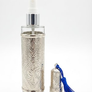 Può includere: Un flacone di profumo in vetro trasparente con un rivestimento in metallo argentato con un design in rilievo ornato. Il flacone ha un nebulizzatore bianco e un tappo argentato abbinato con un nappa blu. Il flacone è alto circa 13 cm.