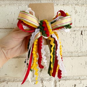 May include: A decorative hair bow featuring a layered arrangement of ribbons in red, yellow, dark green, and white. The design includes various textures such as pom-pom trim, rickrack, and lace, all secured to a brown cardboard display card.