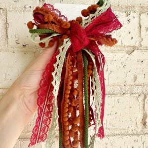 Puede incluir: Un lazo decorativo hecho de una colección de cintas y adornos superpuestos en tonos rojo intenso, verde salvia y naranja quemado. El accesorio presenta varias texturas, incluyendo encaje, flecos de pompones y cinta de grosgrain, atados juntos.