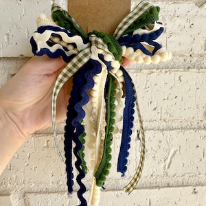 May include: A decorative hair bow featuring a mix of ribbons in navy blue, forest green, cream, and a green and white gingham check pattern. The design includes various textures such as rickrack, pom-pom trim, and flat fabric ribbons.