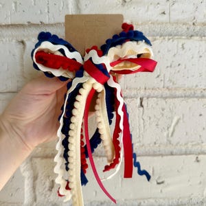 May include: A decorative hair bow featuring layered ribbons in red, white, and navy blue. The design incorporates various textures, including smooth grosgrain, wavy rickrack trim, and a delicate pom-pom fringe, all secured to a brown cardstock backing.