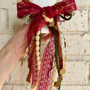 Puede incluir: Un lazo decorativo con encaje rojo oscuro, hilos colgantes de cuentas de madera clara, cordón verde oscuro, ribete de pompones marrones y finas cadenas metálicas doradas con motivos florales sobre una tarjeta rectangular blanca.
