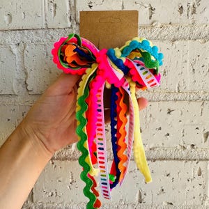 May include: A multi-coloured hair bow featuring a variety of ribbons including neon pink, bright yellow, royal blue, and green rickrack trim. The bow is attached to a brown cardboard display card and includes a white ribbon with a crayon print.