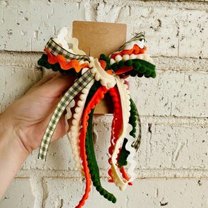 May include: A decorative bow made from multiple layered ribbons in shades of orange, dark green, cream, and a green and white gingham check pattern. The bow features various textures including pom-pom trim and wavy rickrack, attached to a brown card.