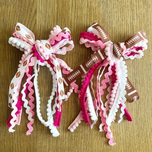 May include: Two decorative hair bows featuring a layered design of ribbons and trim. One bow has a white base with brown football prints, while the other has a brown base with white football prints. Both include pink and white rickrack and pom-pom trim.
