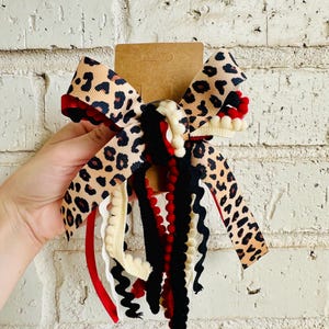 May include: A hair bow featuring a leopard print ribbon layered with black, red, and cream-colored pom-pom trim and rickrack ribbon. The bow is attached to a brown cardboard display card, showcasing a mix of textures and patterns in a multi-layered design.