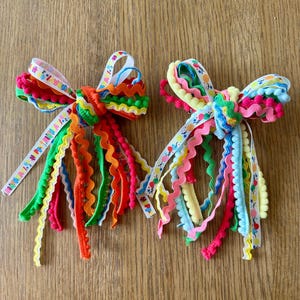 Puede incluir: Dos lazos para el pelo hechos de cintas superpuestas en colores vibrantes como rojo, naranja, verde, azul, rosa y amarillo. Las cintas incluyen ribetes en zigzag, flecos de pompones y una cinta blanca estampada con globos de colores y el texto HAPPY.