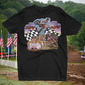 May include: Black t-shirt featuring a motocross design. The graphic includes two racers, checkered flags, and the text "AF7". A portrait of a racer wearing a "THOR" shirt is also present. American flags are visible in the background.
