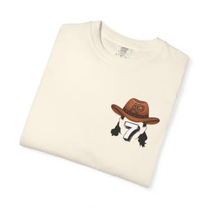 May include: Cream-colored t-shirt featuring a graphic of a brown cowboy hat with the initials "AP" and the number "7". The design includes black hair and is printed on the front of the shirt. The shirt is folded.