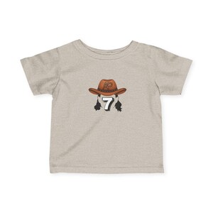 May include: A light gray t-shirt featuring a brown cowboy hat with the letters "AP" on the front. Below the hat is the number "7" with black hair on each side. The shirt is a short-sleeved crew neck.