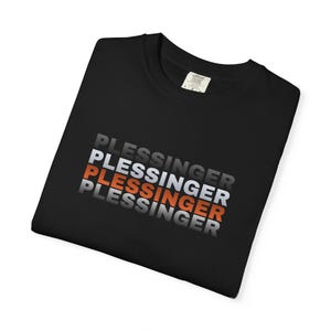 May include: Black t-shirt with the word "PLESSINGER" stacked in a repeating pattern. The text alternates between white, orange, and gray. The shirt is folded and has a tag in the upper right corner.