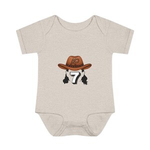 May include: A light beige baby bodysuit with a cowboy-themed graphic. The graphic features a brown cowboy hat with the letters "AP" on it, a white number "7", and black hair. The bodysuit is made of soft material.