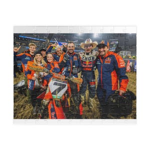 May include: A puzzle featuring a group of people in racing attire, posing for a photo. The group is composed of men and women, some holding trophies and wearing jackets with logos. The background shows a dirt track.