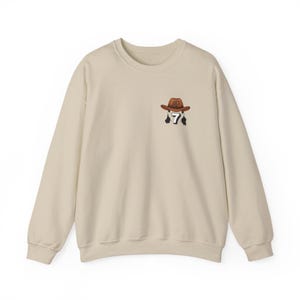 May include: Beige crewneck sweatshirt with a small embroidered design. The design features a brown cowboy hat with the number 7 and two black feathers. The sweatshirt is a neutral color, suitable for various outfits.