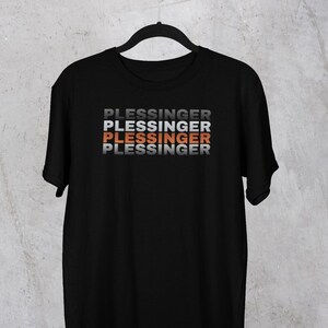 May include: Black t-shirt with the name "PLESSINGER" printed in a stacked design. The text alternates between gray, white, and orange. The shirt is hanging on a black hanger against a neutral background.
