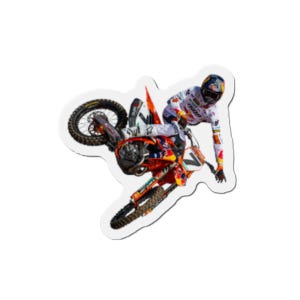 May include: A sticker featuring a motocross rider mid-air on an orange and black motorcycle. The rider wears a white and red racing suit and a helmet. The motorcycle has the number 7 on it.