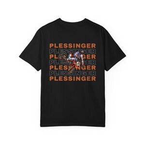 May include: Black t-shirt featuring a motocross rider in action. The shirt has the name "PLESSINGER" stacked vertically in orange and white. The design showcases a dynamic image of a rider on a motorcycle.