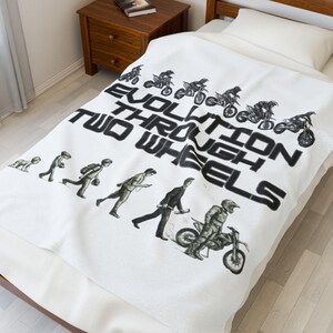 May include: White blanket with the text "EVOLUTION THROUGH TWO WHEELS" in black. The blanket features a graphic of the evolution of man to a motocross rider. The blanket is on a bed with a wooden frame.