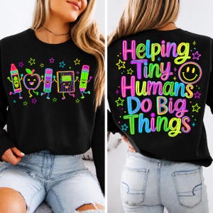 May include: A black long-sleeve sweatshirt featuring a front graphic of colorful cartoon school supplies including a crayon, apple, glue stick, notebook, and pencil. The back displays the text Helping Tiny Humans Do Big Things in neon colors.