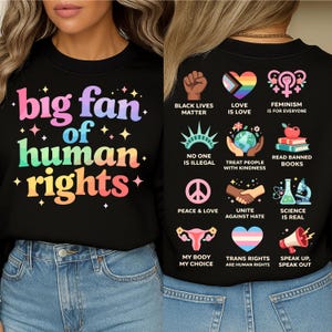 May include: Black sweatshirt with the text "big fan of human rights" in colorful letters. The back of the shirt features various graphics and phrases, including "Black Lives Matter," "Love is Love," and "Trans Rights are Human Rights."