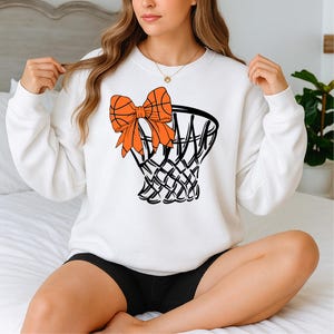 Puede incluir: Sudadera blanca con un dise&ntilde;o de red de baloncesto con un lazo y una pelota de baloncesto de color naranja. El gr&aacute;fico es en negro y naranja. La sudadera es de cuello redondo y manga larga.