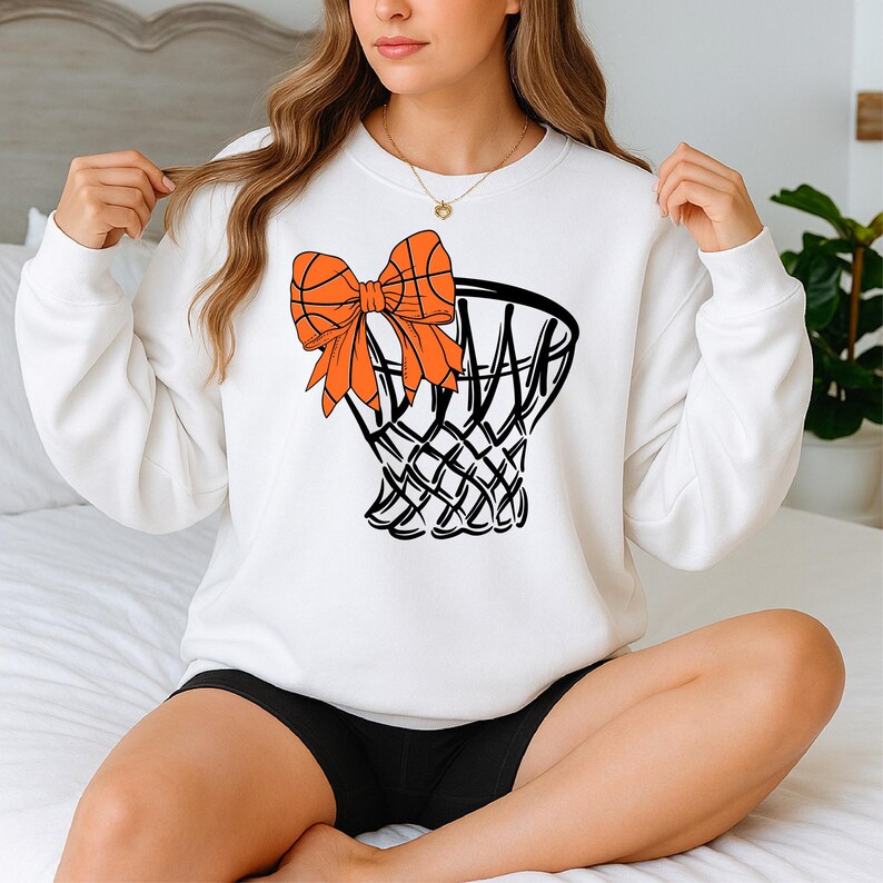 Puede incluir: Sudadera blanca con un dise&ntilde;o de red de baloncesto con un lazo y una pelota de baloncesto de color naranja. El gr&aacute;fico es en negro y naranja. La sudadera es de cuello redondo y manga larga.