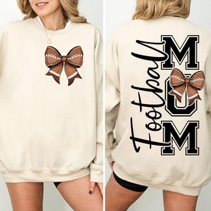 May include: A cream-colored sweatshirt featuring a football-shaped bow design on the front. The back of the sweatshirt displays the words "Football Mom" in black script and block letters, with a football bow graphic.