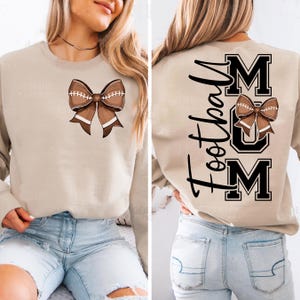 May include: Beige sweatshirt featuring a brown football bow graphic on the front. The back of the sweatshirt displays the word "Football" in black script, along with the word "MUM" in bold black letters, and a football bow graphic.