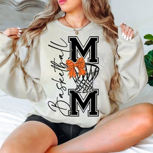 Puede incluir: Sudadera color crema con la frase "Basketball Mom" en escritura negra. El dise&ntilde;o incluye la palabra "MOM" en letras negras en negrita, una red de baloncesto y un lazo naranja. La sudadera es de estilo casual y c&oacute;modo.