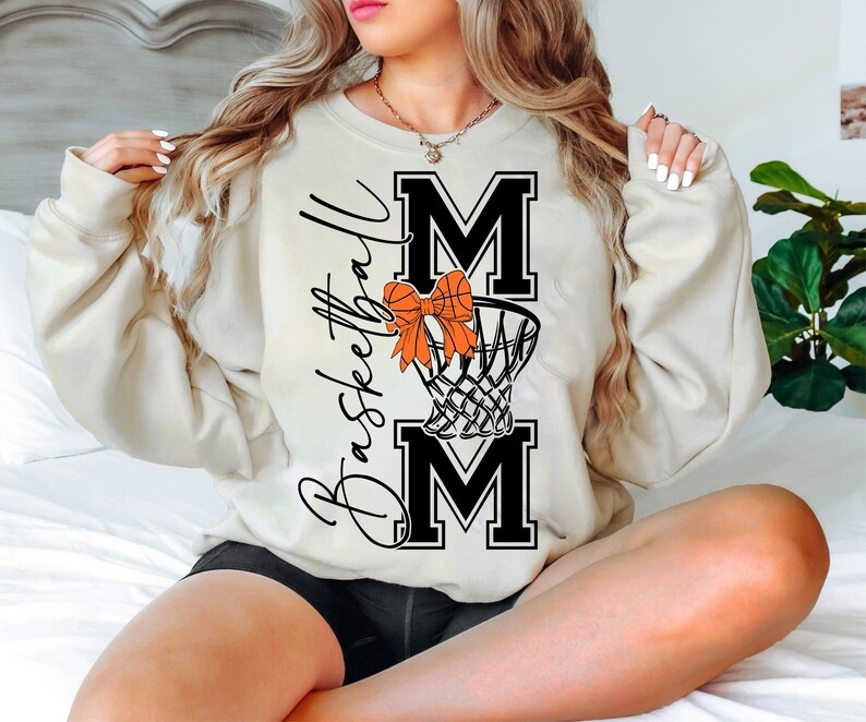 Puede incluir: Sudadera color crema con la frase "Basketball Mom" en escritura negra. El dise&ntilde;o incluye la palabra "MOM" en letras negras en negrita, una red de baloncesto y un lazo naranja. La sudadera es de estilo casual y c&oacute;modo.