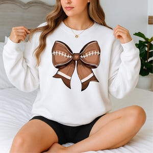 May include: White sweatshirt featuring a large brown bow design. The bow has a football-like appearance with white stitching and brown and white stripes. The sweatshirt is a casual, comfortable style.