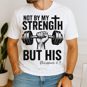 May include: White t-shirt with a black graphic of a hand lifting a barbell, a cross, and the text "NOT BY MY STRENGTH BUT HIS Philippians 4:13". The shirt is made of a soft material.