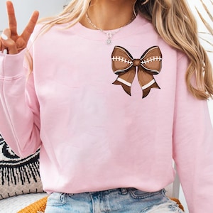 May include: A light pink sweatshirt featuring a brown football-shaped bow graphic. The bow has white stitching and white stripes. The sweatshirt is made of a soft material and has long sleeves.
