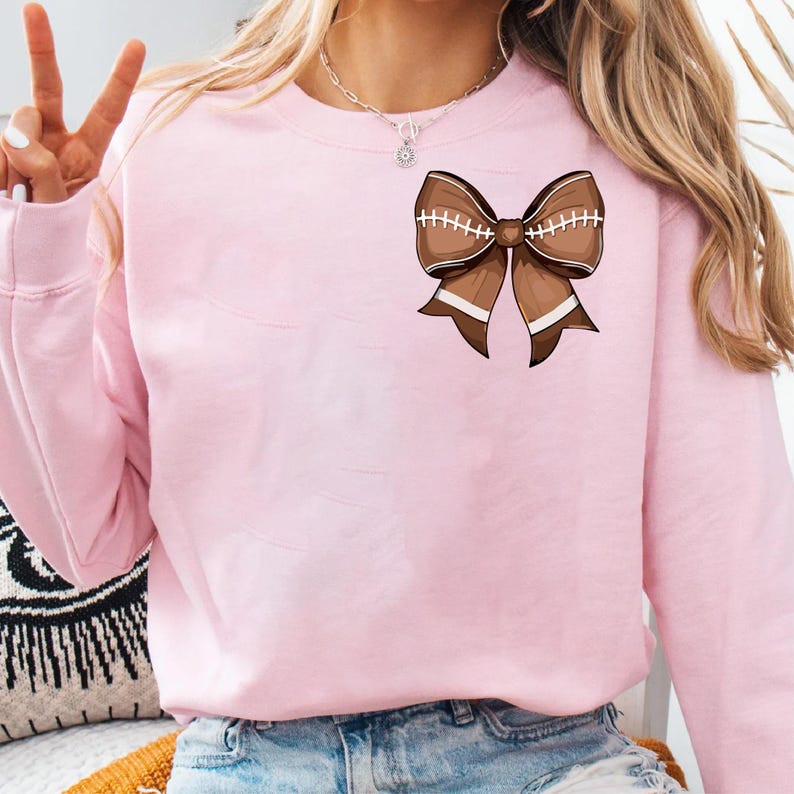 May include: A light pink sweatshirt featuring a brown football-shaped bow graphic. The bow has white stitching and white stripes. The sweatshirt is made of a soft material and has long sleeves.