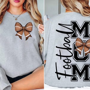 May include: A light gray sweatshirt featuring a brown football-shaped bow. The back of the sweatshirt displays the words "Football Mum" in black. The bow design is repeated on the back, above the word "MUM".