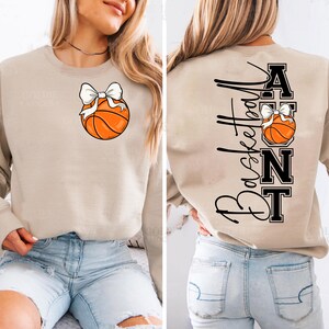 Op de afbeelding: Een beige sweatshirt met een basketbal en een witte strik. De achterkant van de sweatshirt heeft verticaal het woord "Basketball" en het woord "Aunt" met een basketbal- en strikdesign.