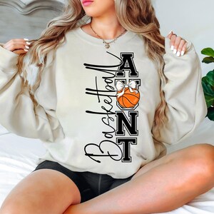 Op de afbeelding: Cr&egrave;mekleurige sweatshirt met de tekst "Basketball Aunt" in zwart script en blokletters. Een basketbal met een witte strik is in het ontwerp verwerkt. De sweatshirt is een casual, comfortabele stijl.
