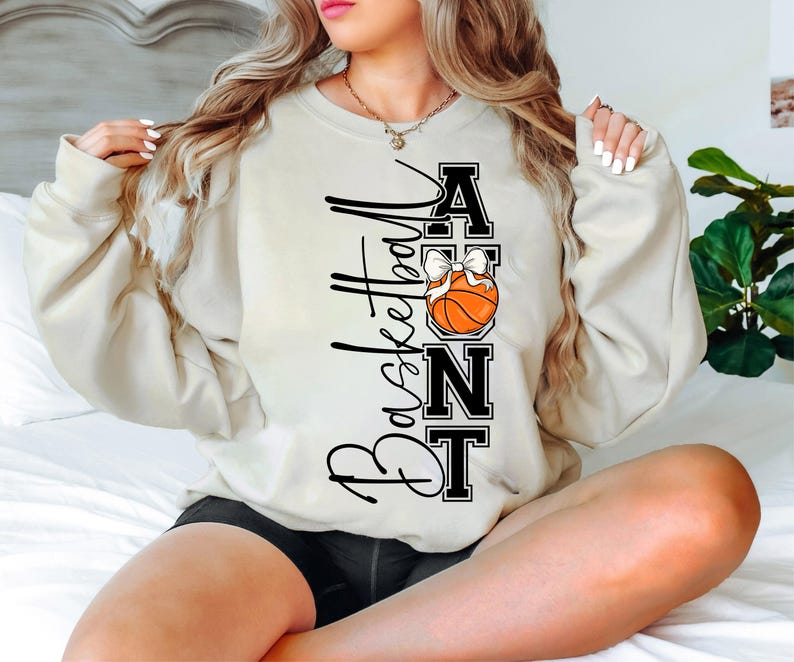 Op de afbeelding: Cr&egrave;mekleurige sweatshirt met de tekst "Basketball Aunt" in zwart script en blokletters. Een basketbal met een witte strik is in het ontwerp verwerkt. De sweatshirt is een casual, comfortabele stijl.