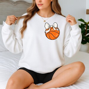 Op de afbeelding: Witte sweatshirt met een basketbalontwerp en een witte strik. De basketbal is oranje met zwarte lijnen. De sweatshirt heeft lange mouwen en een ronde hals.