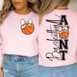 Op de afbeelding: Lichtroze sweatshirt met een basketbal en witte strik. De achterkant toont het woord "Basketball" in zwart script en het woord "Aunt" in blokletters.