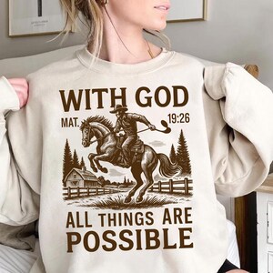 Op de afbeelding: Beige sweatshirt met een bruine afbeelding van een cowboy te paard, met de tekst "WITH GOD MAT. 19:26 ALL THINGS ARE POSSIBLE". Het ontwerp bevat een schuur, bomen en een hek.