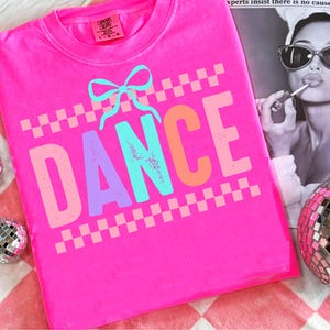May include: A vibrant pink t-shirt featuring the word "DANCE" in multi-coloured letters and a light blue bow. A checkered pattern frames the word. Made from Comfort Colors material.
