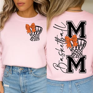 Puede incluir: Sudadera rosa claro con un dise&ntilde;o de red de baloncesto y lazo. La parte trasera presenta la palabra "Basketball" en cursiva, con "MOM" en letras negritas, y un dise&ntilde;o de red de baloncesto y lazo.