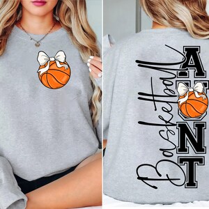 Op de afbeelding: Een lichtgrijs sweatshirt met een basketbal en een witte strik. De achterkant van de trui heeft de woorden "Basketball Aunt" in zwart script en blokletters, met een basketbal en strik ontwerp.