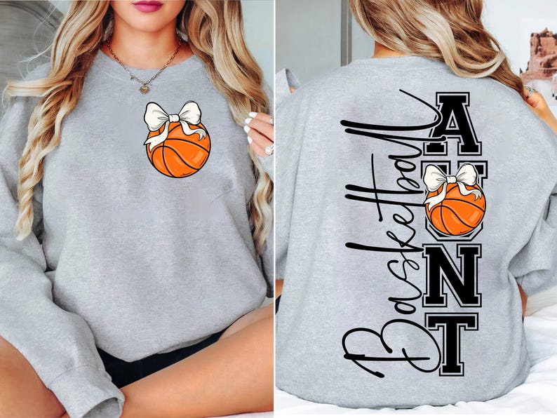 Op de afbeelding: Een lichtgrijs sweatshirt met een basketbal en een witte strik. De achterkant van de trui heeft de woorden "Basketball Aunt" in zwart script en blokletters, met een basketbal en strik ontwerp.