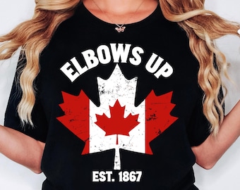 May include: Black t-shirt with a white distressed Canadian maple leaf design. The text "Elbows Up" is above the leaf and "Est. 1867" is below the leaf.